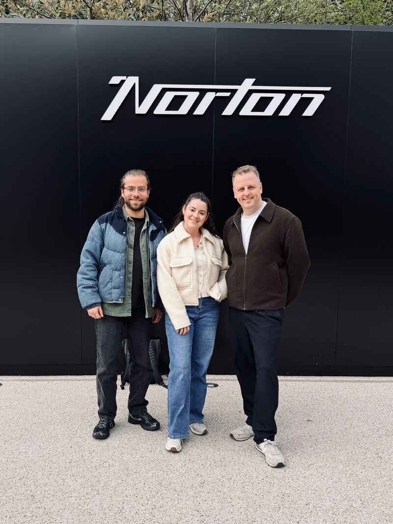 Three people posing in front of a black Norton sign.